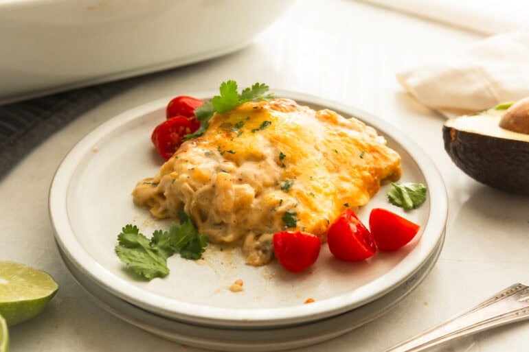 a serving of creamy chicken enchilada casserole on a dinner plate with cilantro sprinkled around it and halved cherry tomatoes around it