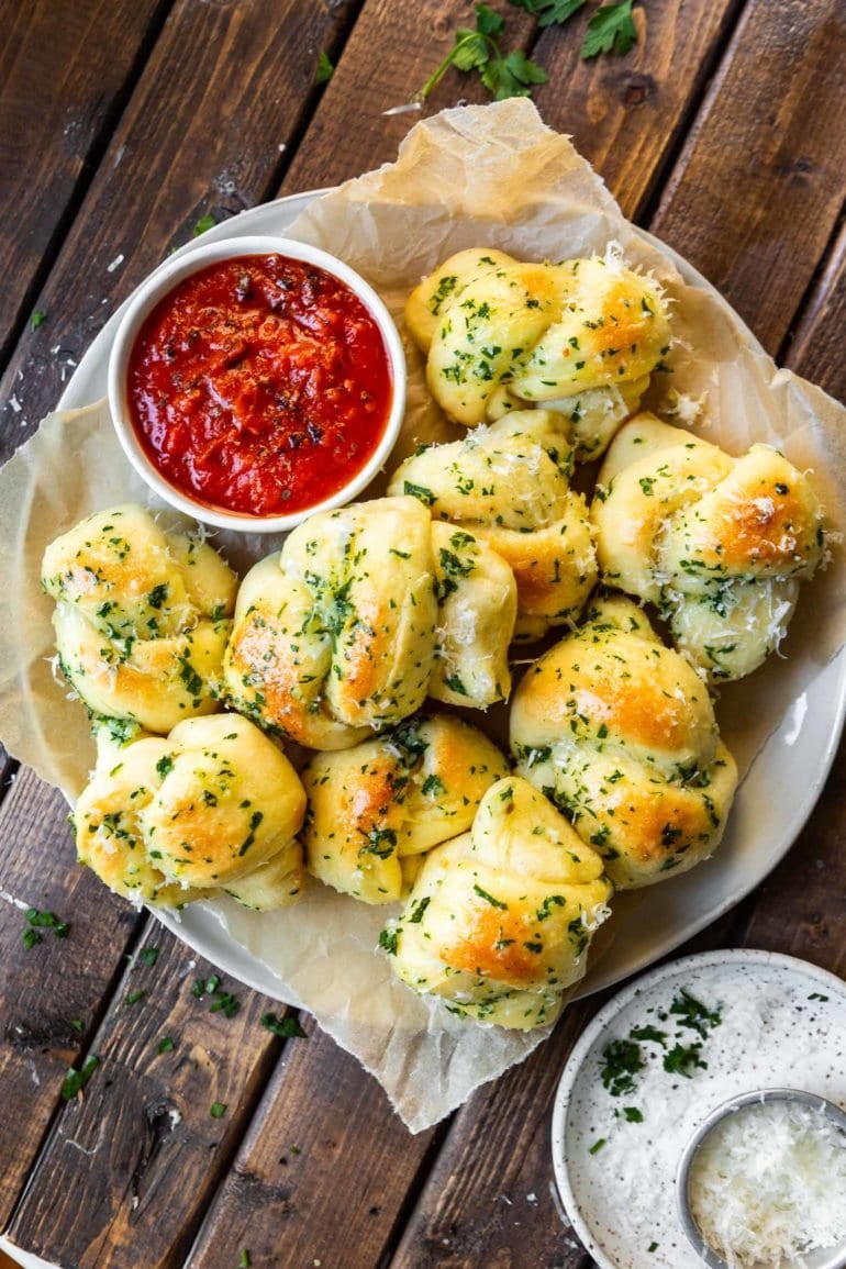 a photo of a plate of italian bread bites topped with melted butter, fresh chopped parsley and parmesan cheese. There is a cup of pizza sauce on the side of the plate.