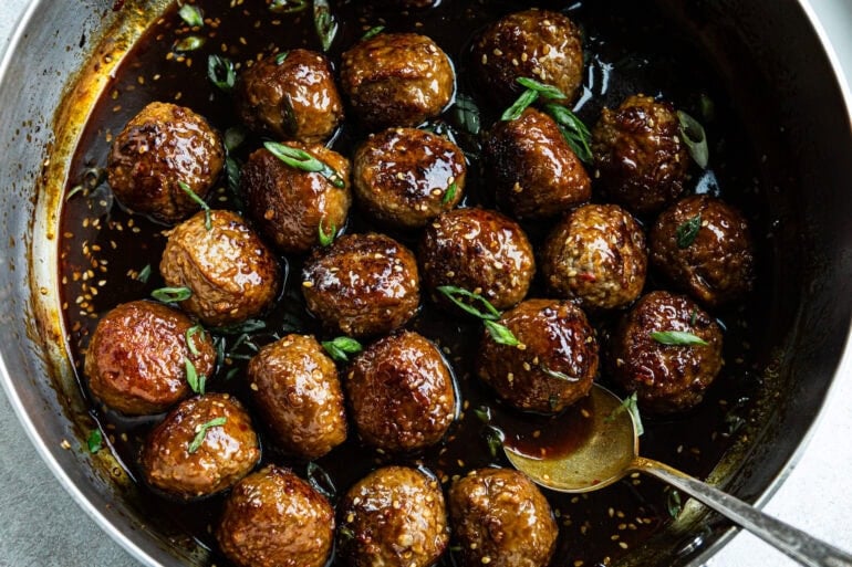 a large skillet full of browned meatballs in a sticky sauce and topped with chopped green onions and sesame seeds