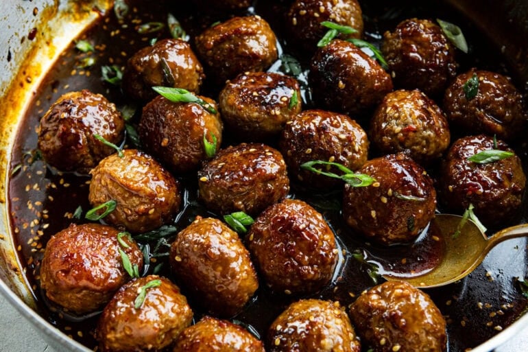 several golden brown meatballs sitting a thick stick sauce and topped with sesame seeds and green onions