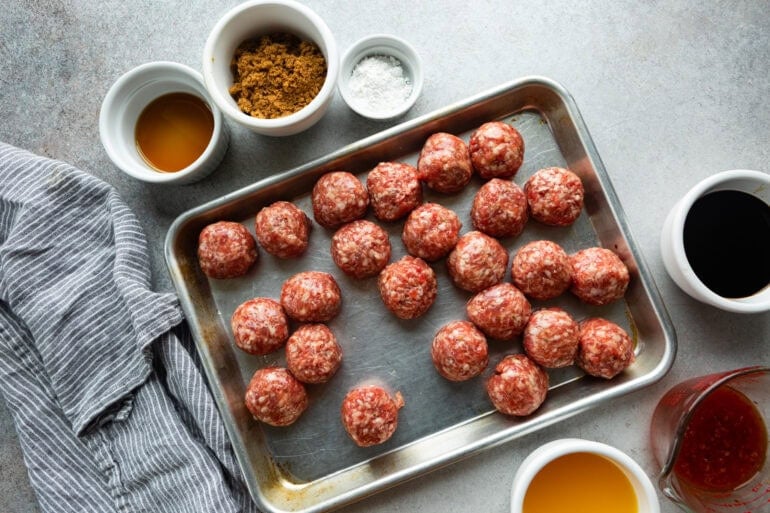 all the ingredients for sticky korean meatballs with raw meatballs on a baking sheet, brown sugar, oil, salt, etc all in separate small bowls