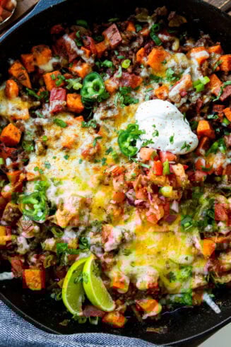 a photo of a cast iron skillet full of taco ground beef and diced sweet potatoes topped with a dollop of sour cream, chopped fresh cilantro and lime wedges