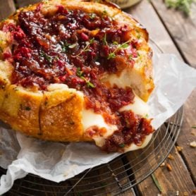 A bread bowl filled with brie and topped with onion jam and raspberries. It has been baked until it is golden brown and the cheese is melted.