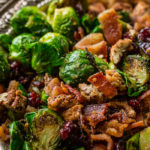 A vintage, silver bowl with flower full of bright green brussels sprouts that have been halved and pan roasted until golden brown on one side, olive oil and seasoning covering all of them and tossed with red craisins, crisp bacon and crunchy candied pecans ohsweetbasil.com