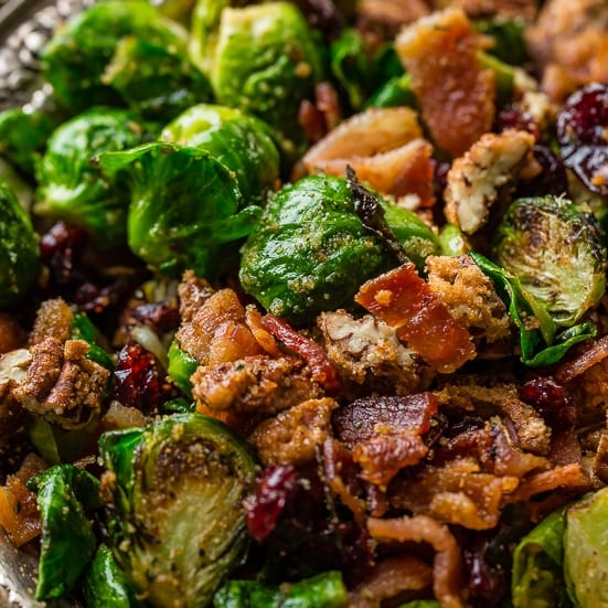 a bowl full of green brussels sprouts that have been roasted in a pan and are golden brown, sprinkled with seasoning and tossed with candied pecans, bacon pieces and red craisins as a side dish for any meal