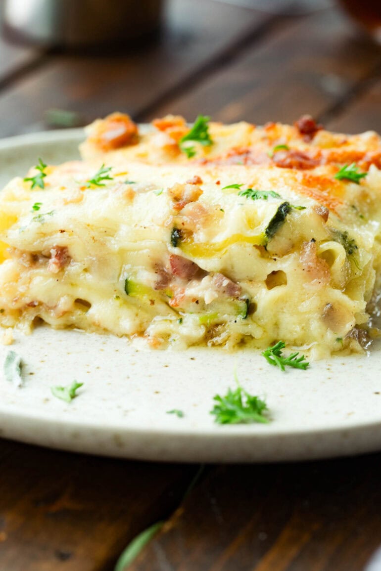 a photo taken from the side of a piece of pancetta vegetable lasagna sitting on a dinner plate so you can see all the cheesy layers.