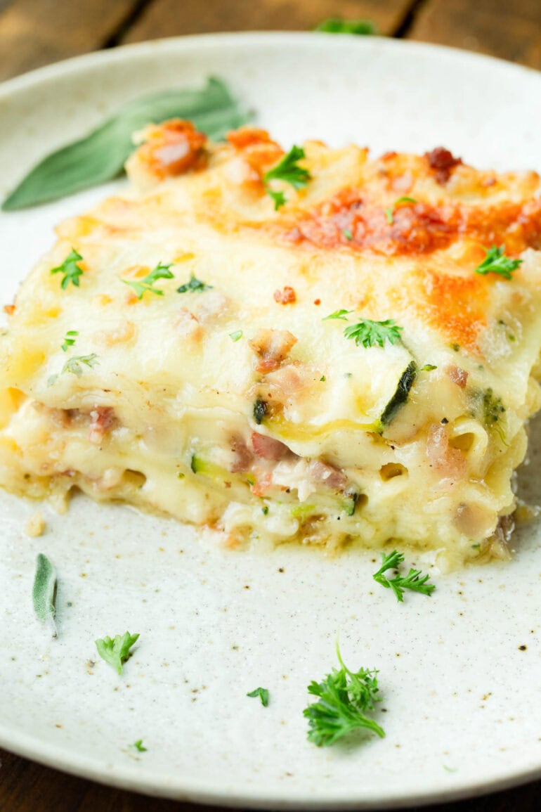 a photo of a serving of pancetta vegetable lasagna with white sauce sitting on a white dinner plate