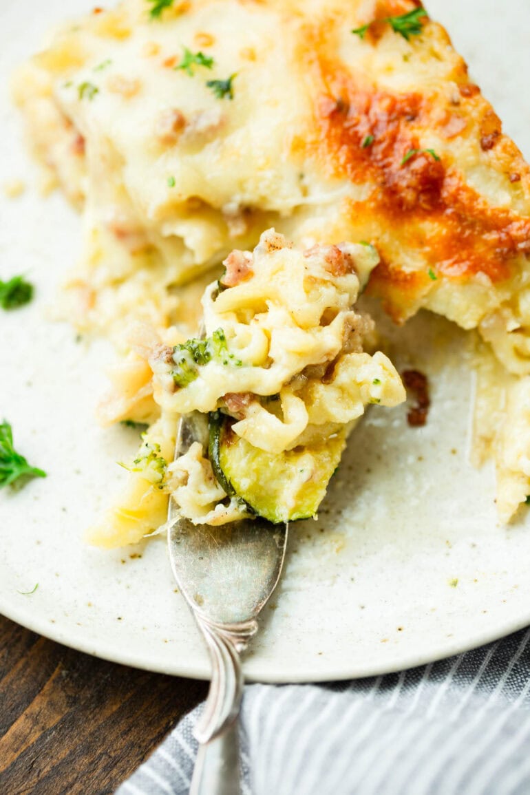 a photo of a piece of pancetta vegetable lasagna with white sauce being cut up and eaten with a fork