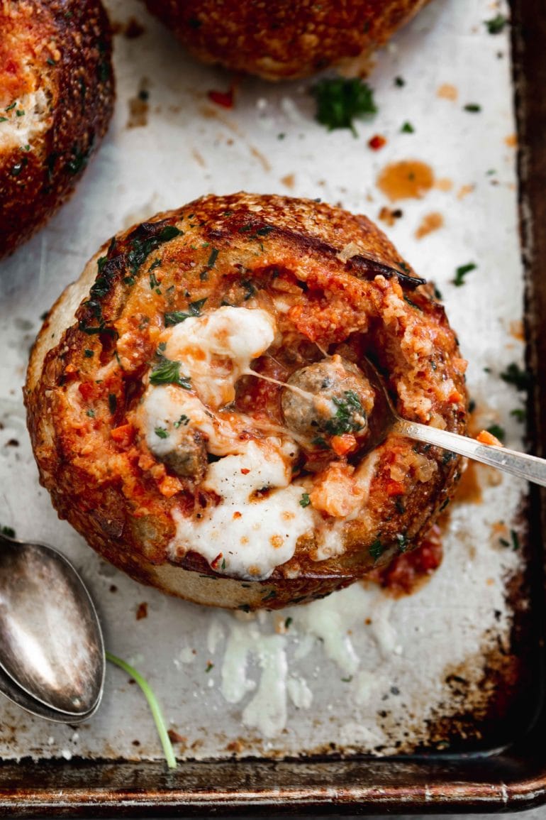 A bread bowl filled with parmesan meatball soup. A spoon is in the soup and cheese is melted on top.