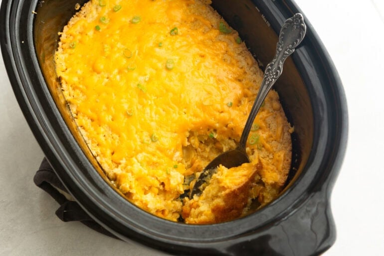 a crock pot full of corn casserole with a large serving spoon scooping up serving
