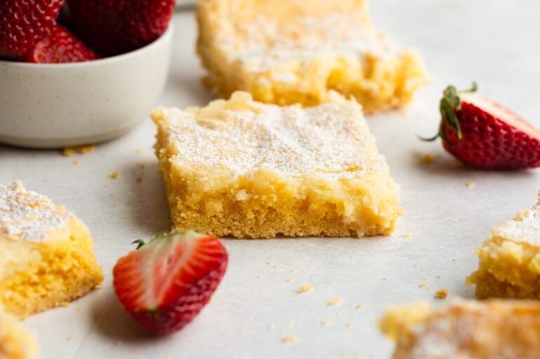 A slice of ooey gooey butter cake surrounded by other cut slices and cut strawberries.