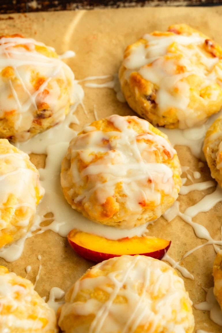 several biscuits on parchment paper with a drizzle of glaze and peach slices on the side.
