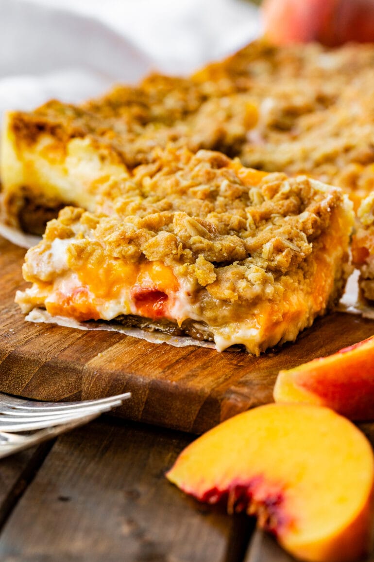 a photo of a peaches and cream crumble bar topped with a golden crumble topping with juicy peaches and creamy filling coming out the sides.