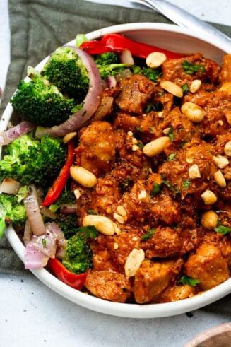 a photo of saucy peanut butter chicken topped with chopped peanuts and a side of broccoli, red onion slices and red pepper slices