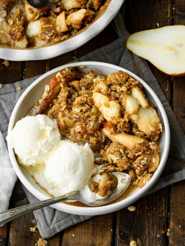 a photo of a bowl of pear crisp with juicy tender pears, topped with crunchy oat topping and two scoops of vanilla ice cream on the side.
