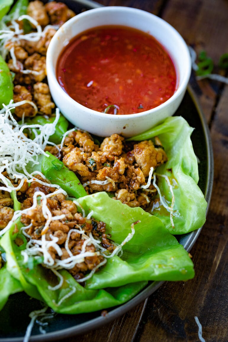 a photo of chicken lettuce wraps topped with crunchy rice noodles sitting next to a cup of sweet chili dipping sauce.