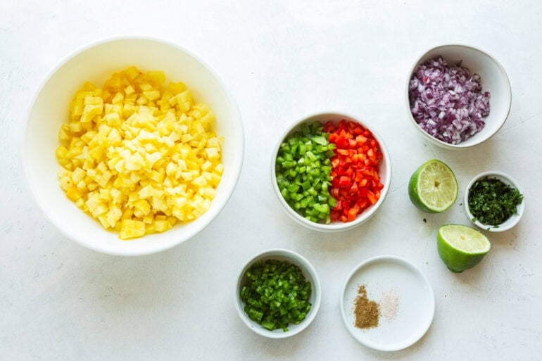 all the ingredients for fresh pineapple salsa in separate small bowls including pineapple, red and green bell pepper, red onion, cumin, jalapeno and salt