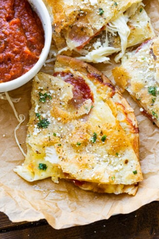 a photo of a slice of a tortilla pizza that is golden and crispy with melty cheese coming out and topped with sprinkled cotija cheese and garlic butter with a small bowl of pizza sauce next to it