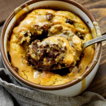 a photo of a mug full of baked oatmeal peanut butter and chocolate mug cake and someone is removing a spoonful of cake from the middle.
