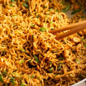 a pair of chopsticks lifting saucy peanut butter ramen from the pan topped with green onions, sesame seeds and chopped peanuts