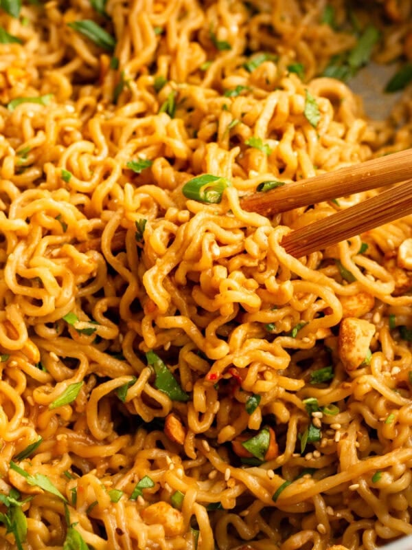 a pair of chopsticks lifting saucy peanut butter ramen from the pan topped with green onions, sesame seeds and chopped peanuts