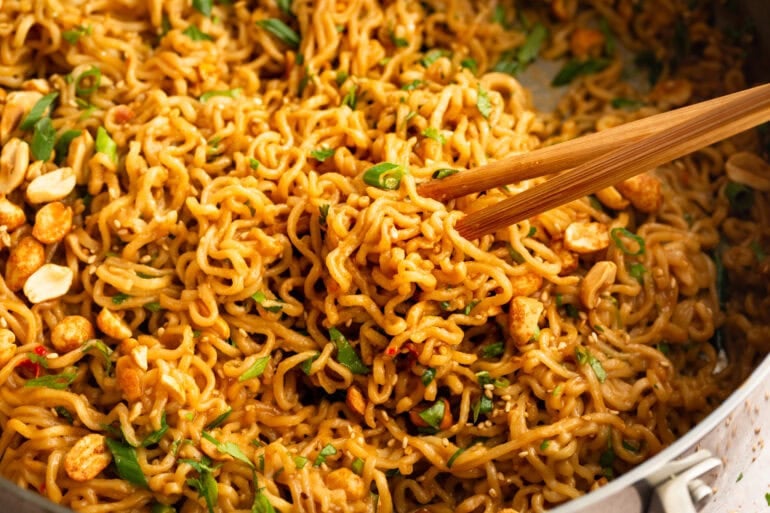 a pair of chopsticks lifting saucy peanut butter ramen from the pan topped with green onions, sesame seeds and chopped peanuts