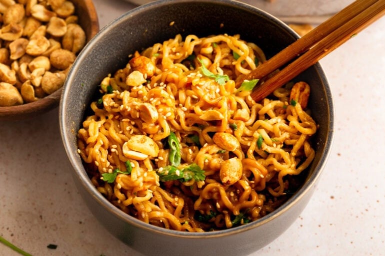 a bowl of peanut butter ramen topped with chopped peanuts, green onions and sesame seeds with chopsticks sticking into the bowl