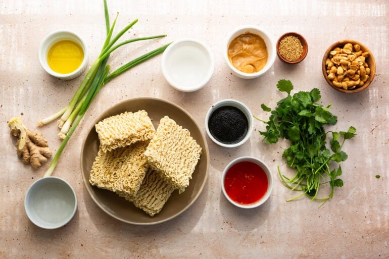 all the ingredients for peanut butter ramen in separate small bowls including dry ramen, creamy peanut butter, sweet chili sauce, soy sauce, ginger, cilantro, etc
