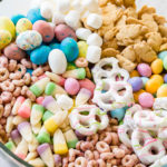 Easter snack mix in a clear glass bowl with berry Cheerios, Easter candy corns, yogurt covered pretzels, Robin eggs, Easter M&Ms. mini marshmallows and bunny cookies.