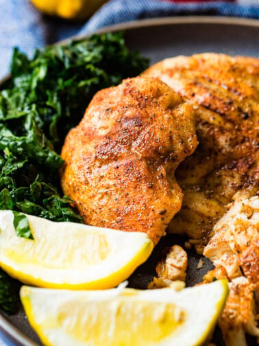 A photo of whole smoked chicken breasts and on a charcoal gray plate with lemon wedges and kale on the side.