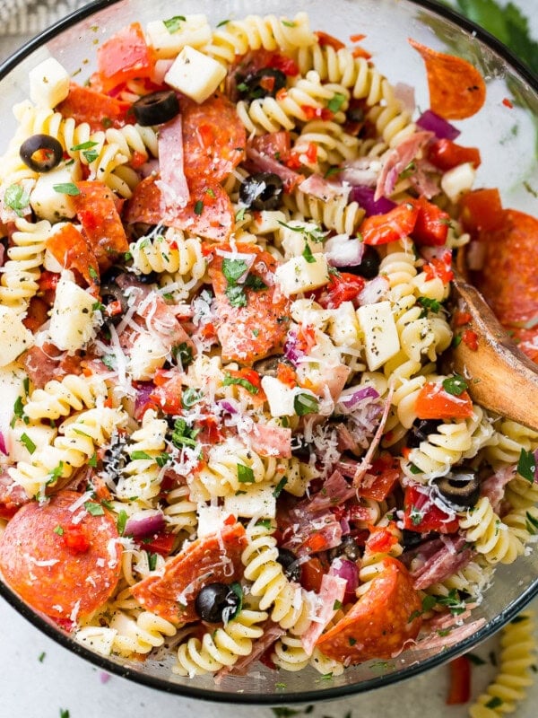 A glass bowl of quick and easy italian pasta salad recipe with homemade italian dressing. Rotini pasta with pepperoni, salsami, black olives, parmesan cheese, mozzarella cheese, peppers, dressing and red onions!