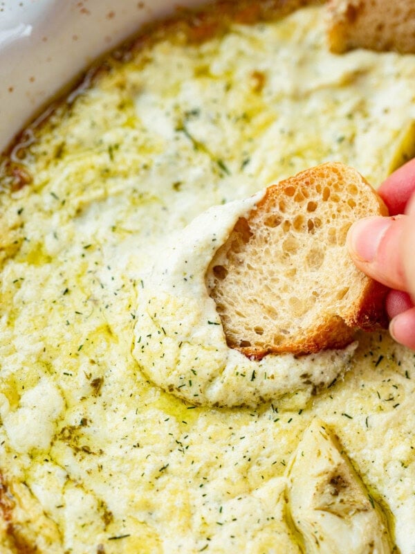 a photo of a hand dipping a crostini in a large dish filled with creamy artichoke dip.