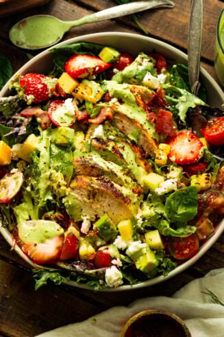 a photo taken over the top of a large serving bowl full of mixed greens, a sliced grilled chicken breast, corn, halved cherry tomatoes all topped with a creamy basil dressing