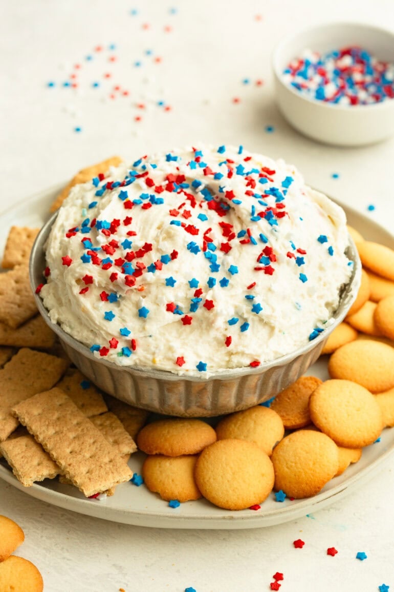 A photo of Funfetti Dip topped with a generous pile of colorful red white and blue sprinkles sitting on a white marble surface next to a stack of graham crackers and nilla wafers.