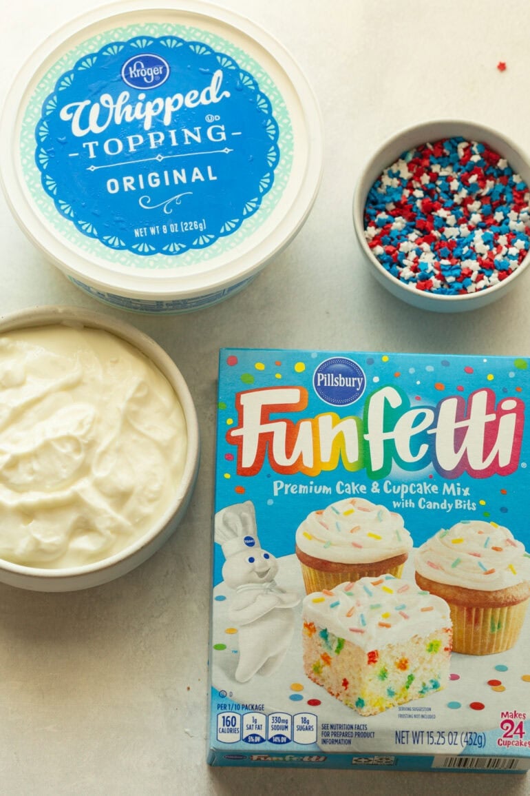 Ingredients like cake mix, sprinkles, and whipped topping for a fluffy creamy cake batter dip topped with red white and blue sprinkles on a Fourth of July party table.