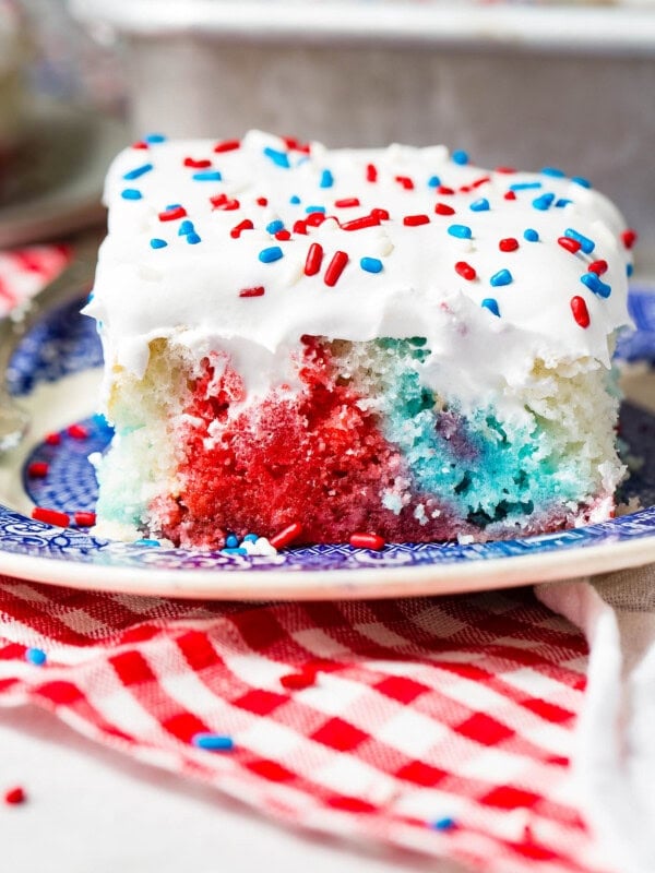 A piece of red, white and blue poke cake on a blue and white dessert plate. The cake is frosted with white frosting and sprinkled with red, white and blue sprinkles. You can see the red, white and blue portions of the cake where holes have been poked and jello poured in. the plate is sitting on a red and white checkered napkin.