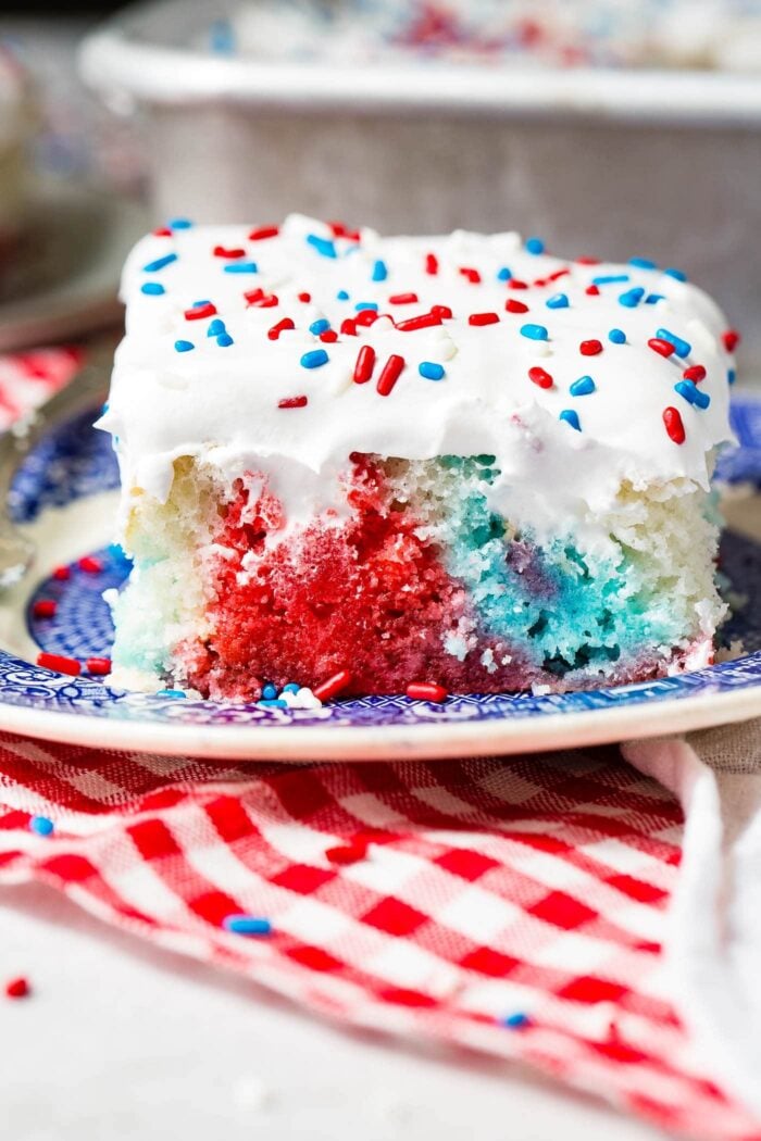 A piece of red, white and blue poke cake on a blue and white dessert plate. The cake is frosted with white frosting and sprinkled with red, white and blue sprinkles. You can see the red, white and blue portions of the cake where holes have been poked and jello poured in. the plate is sitting on a red and white checkered napkin.