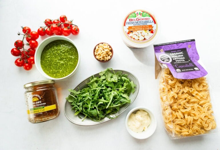 all the ingredients for pesto pasta salad including pasta spirals, pesto, arugula, pine nuts, grape tomatoes, sun dried tomatoes and mozzarella balls