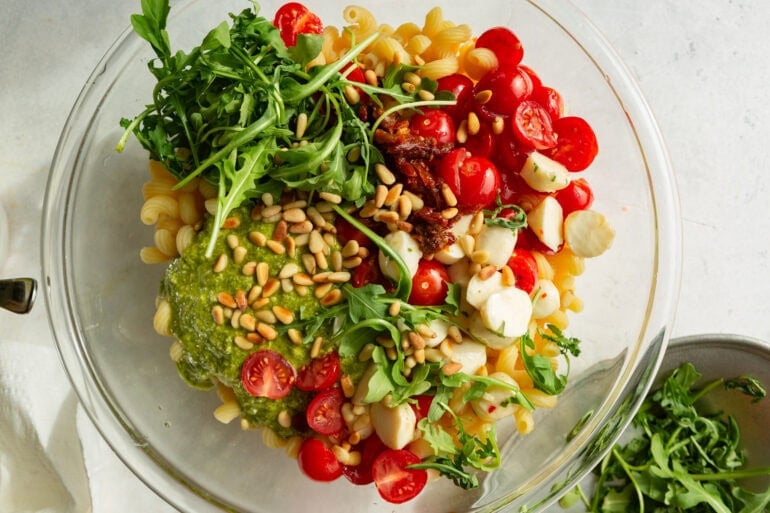 a large glass mixing bowl with all the components of a pesto pasta salad ready to have dressing added and stirred together