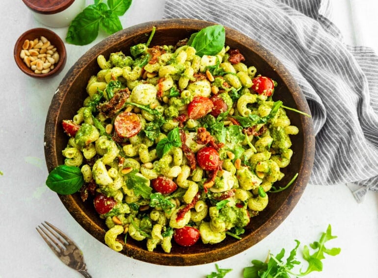a large wooden serving bowl full pesto pasta salad with cherry tomatoes, mozzarella balls, basil leaves and pine nuts