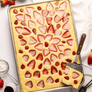 a half sheet pan full of baked sheet pan pancakes topped with slices of fresh strawberry baked into the pancakes and a metal spatula removing one serving