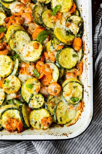 a photo of a white rectangular baking dish full of roasted sliced zucchini, yellow squash and carrots topped with fresh basil and cheese