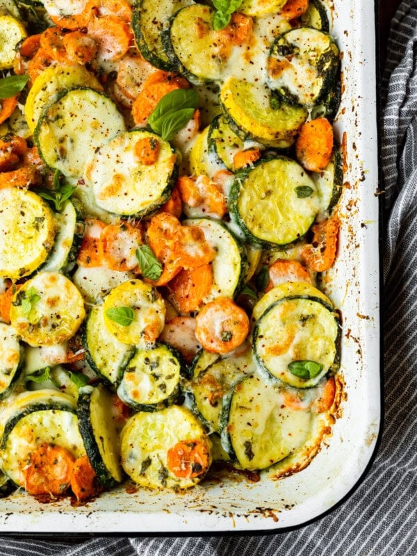 a photo of a white rectangular baking dish full of roasted sliced zucchini, yellow squash and carrots topped with fresh basil and cheese