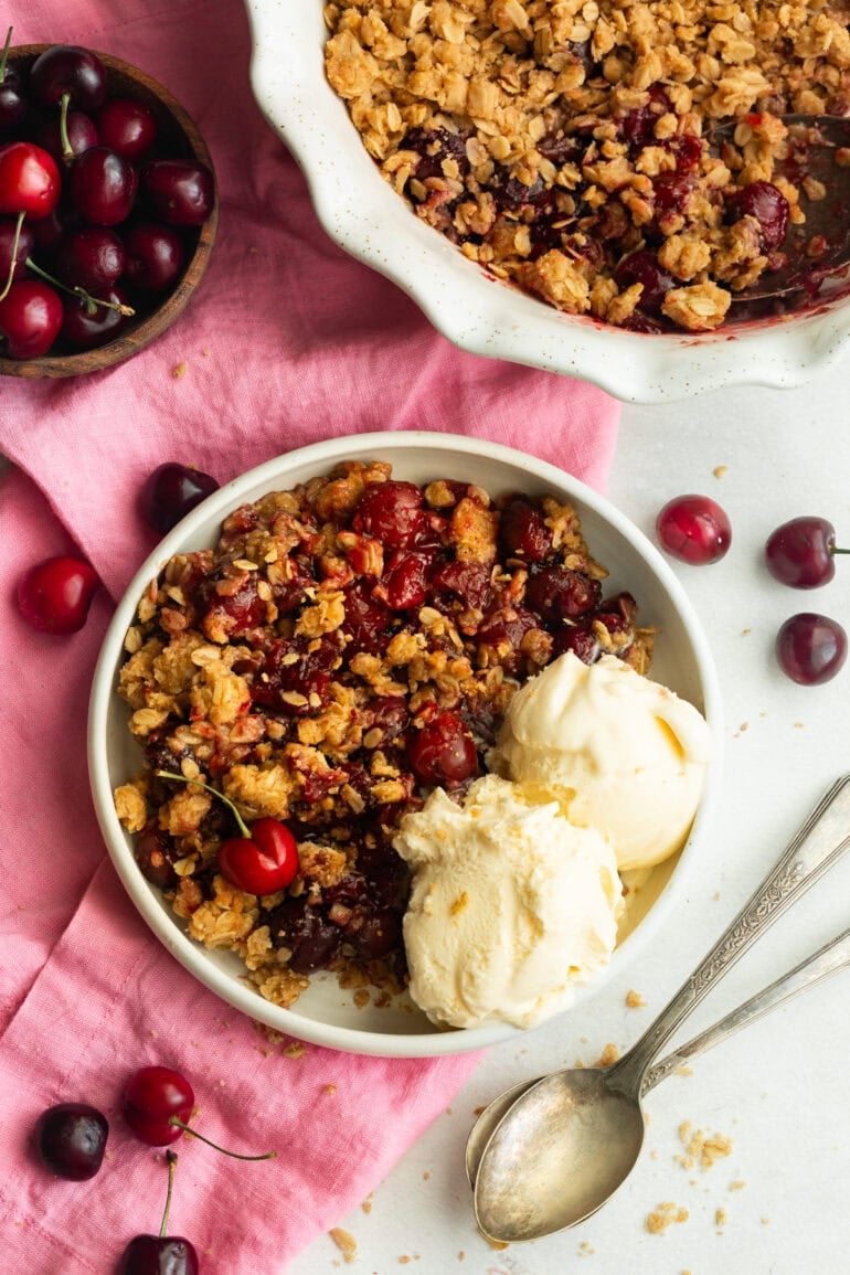 a serving of cherry crisp on a small dessert plate with two scoops of vanilla on the side and the whole cherry crisp in a baking dish sitting next to the plate