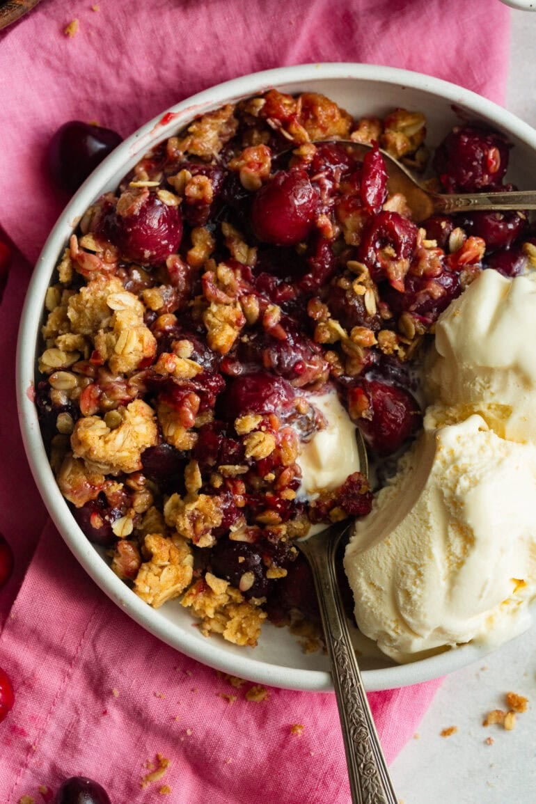 a serving of cherry crisp in a small bowl melting vanilla ice cream on the side of the dish