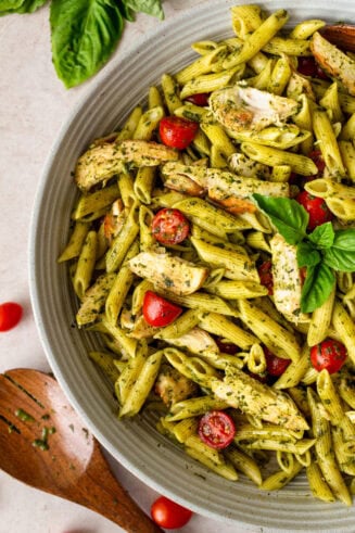 a photo of a large serving bowl full of cooked penne pasta tossed with sliced chicken breasts, sliced grape tomatoes, and fresh basil.