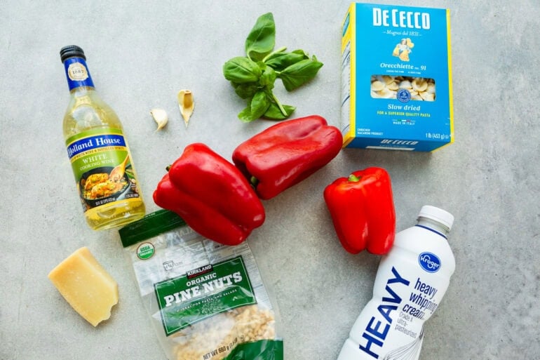 all the ingredients for roasted red pepper pasta sitting on table including red bell peppers, heavy cream, pasta, fresh basil, garlic gloves and parmesan cheese