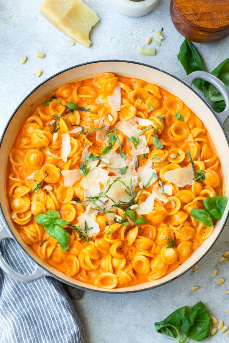 a large dutch oven full of cooker oreicchete pasta mixed in a creamy roasted red pepper sauce and topped with shaved parmesan, chopped basil and toasted pine nuts