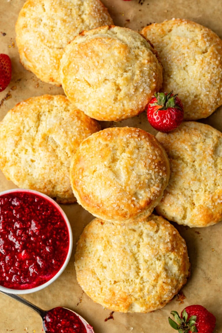 several golden baked sweet cream biscuits sitting on a brown parchment paper with a small bowl of strawberry jam and a whole strawberry sitting with the biscuits