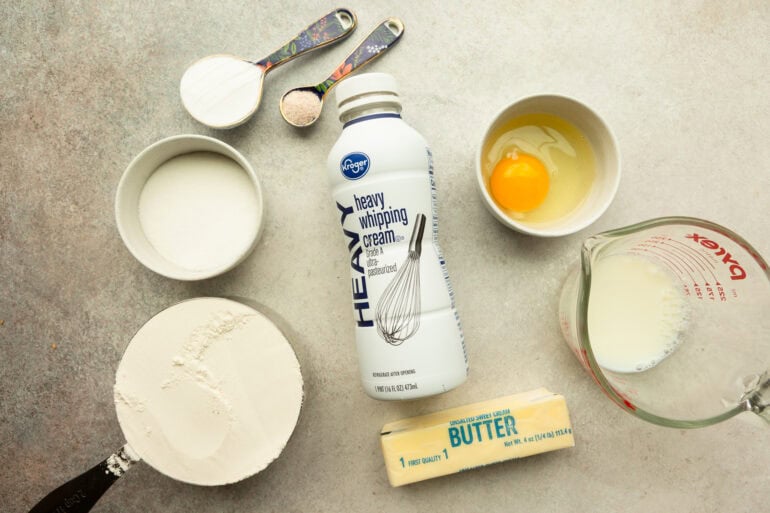 all the ingredients for sweet cream biscuits including a bottle of heavy cream, butter, sugar, flour, and egg and salt